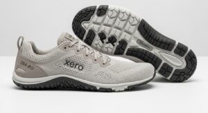 A pair of light grey Xero Shoes featuring branding on the side; one shoe standing upright while the other lies on its side, showcasing a black and white textured sole—engineered for walking and providing all-day comfort with a visible tread pattern.